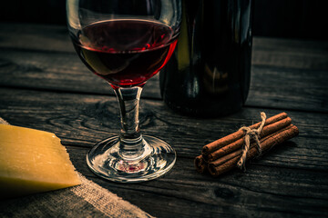 Bottle of red wine with a glass of red wine and a piece of parmesan and cinnamon sticks on an old wooden table. Angle view, focus on the glass of red wine