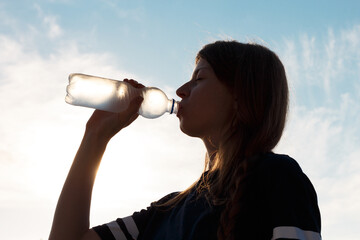 Silhouette of a woman drinking chilled water from a transparent misted plastic bottle against the backdrop of a sunny sky.