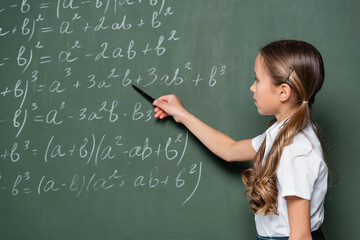 preteen schoolgirl pointing with pen at mathematic equations on chalkboard