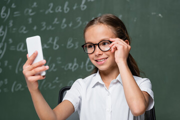 smiling schoolkid touching eyeglasses during video call near chalkboard