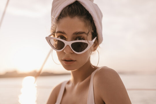 Stylish girl with light hair, blue eyes and thick eyebrows, in sunglasses and towel on hair, looking into camera against sea and sky background during sunset