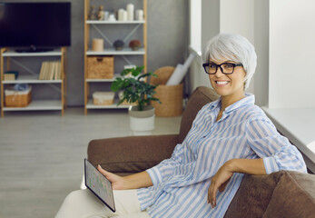 Happy mature woman in glasses enjoying free time on comfortable sofa, holding modern tablet,...