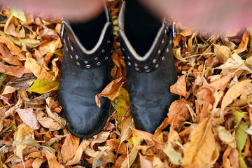 A girl in dirty black boots stands on the autumn leaves. Autumn background in the form of yellow dry leaves and human feet. Dirty shoes due to bad rainy weather in the fall season