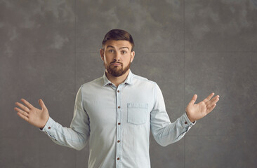 Portrait of a handsome young man standing on a grey studio background shrugging shoulders with a...