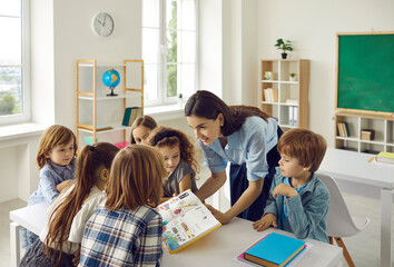 Young friendly caucasian female teacher in the classroom with a book in her hands teaches young students. Modern elementary school students stand around a desk and listen intently to the teacher. - obrazy, fototapety, plakaty