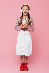 Smiling child in headphones holding milkshake on pink background