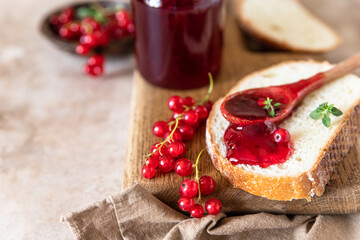 Slice of bread and red currant jam or jelly in a spoon, wooden cutting board.
