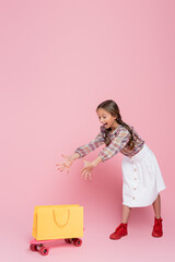 excited girl with open mouth and outstretched hands near yellow shopping bag and penny board on pink background