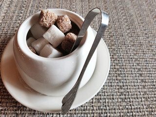 White deep glass bowl with white and dark sugar lumps and metal tweezers on gray backing