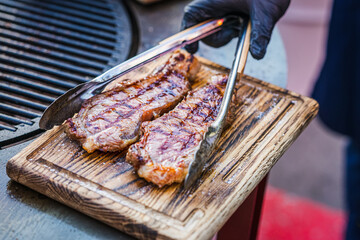 Hand of chef in glove with tongs serving Beef Steaks on wooden cutting board, selective focus