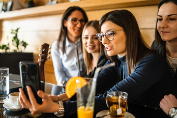 Side view of adult caucasian woman with eyeglasses holding mobile phone while sitting at cafe or restaurant with her female millennial friends making a video call on smartphone or taking selfie photo