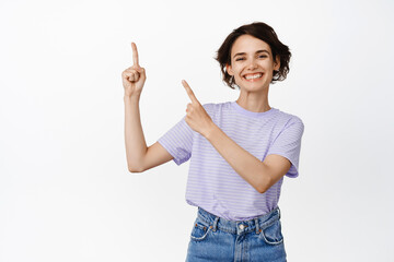 Cheerful brunette girl pointing fingers at upper left corner and smiling proud, showing advertisement, company info, standing in t-shirt against white background