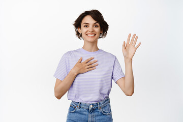 Friendly smiling woman hold arm on heart and palm raised high, making promise, introduce herself, my name is gesture, looking happy, standing over white background