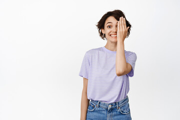 Happy beautiful girl hide one side of face, looking at camera excited, smiling, cover eye, standing in casual clothes against white background