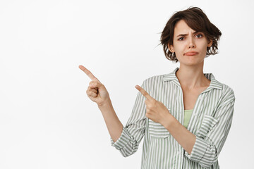 Image of skeptical brunette girl pointing fingers left, showing advertisement and frowning upset,...