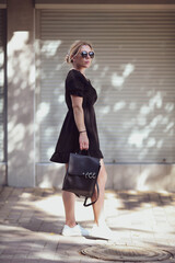 Obraz premium Beautiful blonde with a backpack on her back. Young woman in sunglasses smiles on the background of white roller blinds. Copy space
