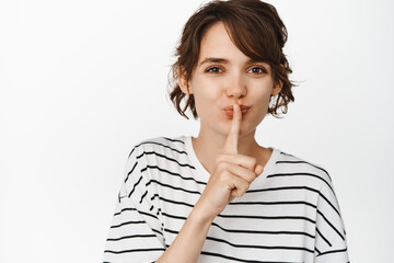 Smiling modern woman showing hush, shush gesture, finger pressed to lips and looking coquettish, hiding secret, standing over white background