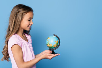Side view of smiling girl holding globe on hands isolated on blue