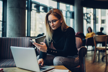 Skilled Caucasian woman in classic spectacles checking received email message on smartphone gadget while sharing media files to netbook using bluetooth connection in coworking, distance job