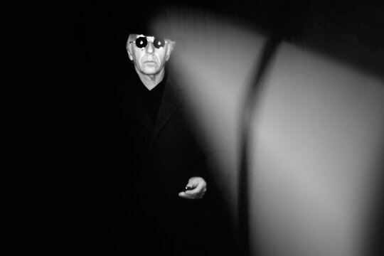Spooky image of a man in black, wearing fedora hat, long coat and sunglasses in a dark place.