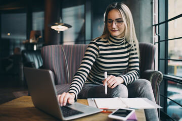 Caucaisan female student with education textbook searching information on netbook while studying, skilled hipster girl in optical eyewear e learning in coworking space using 4g wireless on laptop