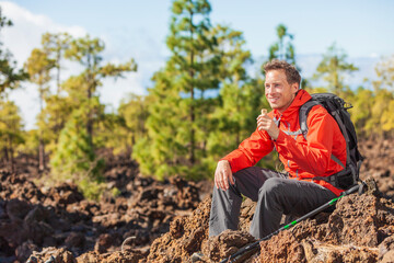 Naklejka na ściany i meble Hiking man eating granola protein bar snack during mountain hike travel camping vacation in nature landscape. Happy hiker taking a break with healthy food in his backpack.