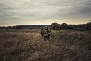 Hunters walking through rural field towards forest against sunrise sky and forest on horizon during...