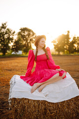 Fototapeta premium Beautiful curly woman in hat and clothes posing near hay bales in the countryside at sunset. Fashion concept. Nature, vacation, relax and lifestyle.