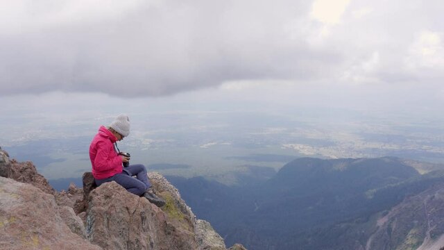 A female hiker sitting on top of a rocky mountain and looking through the photos taken on her camera