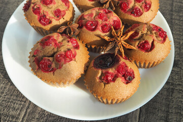 nearby, several gluten-free chocolate muffins with red currants lie on a white round plate with a brown star of anise on a wooden table background. side view . dietary gluten-free cupcake, prepared at