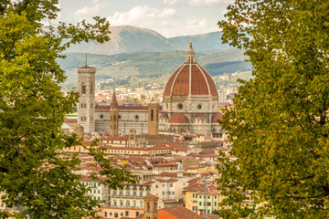 Fototapeta na wymiar Florence's beautiful Gothic cathedral standing out over the surrounding city buildings, with the mountains in the background, framed by some green foliage
