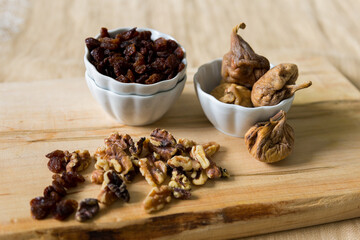 High angle selective focus still life of dried figs and raisins in small white bowls, as well as walnuts against a wooden cutting board background