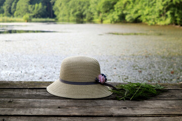 The concept of summer walks in nature. A light hat, a bouquet of ferns on a wooden bridge against the background of a lake, close-up.