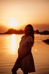 Young woman at sunset. Silhouette of pregnant happy woman with long hair in rays of setting sun, standing on beach and enjoying summer landscape. 