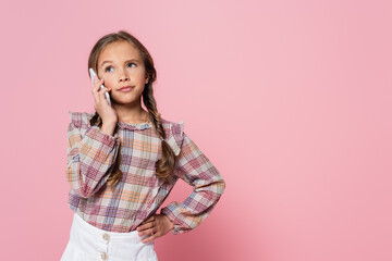 serious preteen girl talking on smartphone while standing with hand on hip isolated on pink