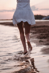 Close-up of a running woman. Photo of legs back view of a beautiful tanned woman in a white summer dress on the river bank against the background of sunset and clouds.