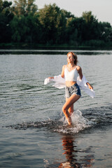 A beautiful young woman runs freedom through the water. A young blonde in a white top and shirt, denim shorts against the background of trees and a river. 