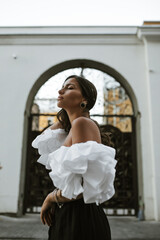 Attractive young tanned brunette in white top with puffed sleeves, black pants and stylish vintage accessories standing sideways outdoors and closing eyes