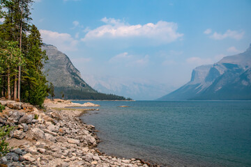 Trees stand back from the glacial water on the rock-lined shore of Lake Minnewanka in Banff...