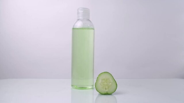 A woman's hand puts, takes on a white table a transparent bottle with a refreshing face tonic from a cucumber, in the foreground - a slice of cucumber