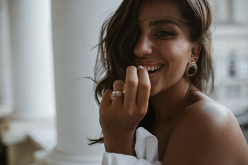 Portrait of young attractive brunette with long hair, smooth tanned body, dark eyes in vintage earrings, ring, looking into camera and smiling against light background