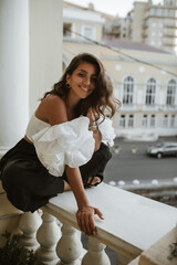 Gorgeous young dark-haired and tanned lady, wearing white vintage top, black pants and stylish accessories, resting at balcony. Great classic architecture, column and city view.