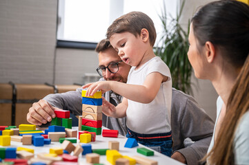 Young happy family. Mother and father playing with her cute toddler son at home using didactic wooden toys. Home education - obrazy, fototapety, plakaty