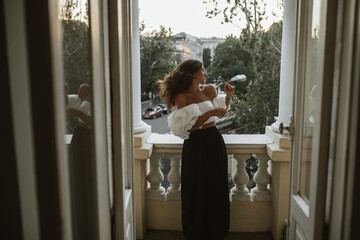 Tanned young brunette in white top with puffed sleeves, black high-wasted pants, smiling and looking at evening city from classic balcony