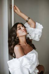 Fototapeta premium Beautiful young lady with curly brunette hair and stylish make-up, in white off-shoulder top, vintage accessories sensually posing indoors and closing eyes