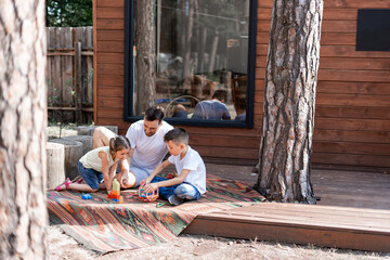 A father plays with two children sitting on the porch near a country wooden house, spends his free time helping children play with toys, spends vacation with his family