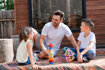 Happy dad or babysitter has fun with little girl and boy, plays with them in the yard sitting on...