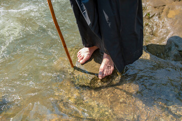 A monk with bare feet stands on the rocks by a mountain river with a staff. Washing the feet of travelers, Bible stories.