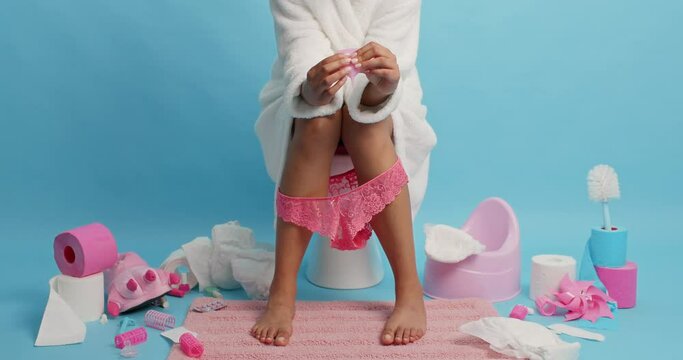 Faceless female model wears white bathrobe lacy panties pulled down on legs holds menstruation cup has monthly periods sits on toilet bowl in lavatory against blue background. Women hygiene.