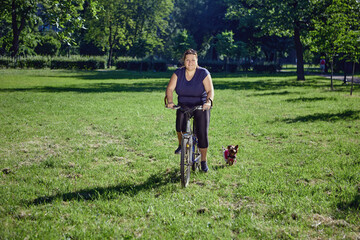 Body positive woman rides bicycle in garden with yorkshire terrier.